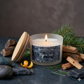 high-end minimalist luxury aesthetic photo of a white frosted glass 'Black Ice' candle with a natural wood lid and a realistic glowing flame. The creamy white wax is showcased in a sophisticated masculine setting on a dark slate surface. Fragrance cues include prominent natural sandalwood blocks, a crisp green pine branch, and a single twist of orange zest, accented by smooth dark river stones and charcoal slate shards against a moody, textured backdrop