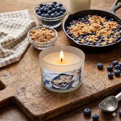 Lit jar candle with "Blueberry Cobbler" label on a rustic wood board next to a fresh blueberry cobbler, bowls of berries, and an ice cream scoop.