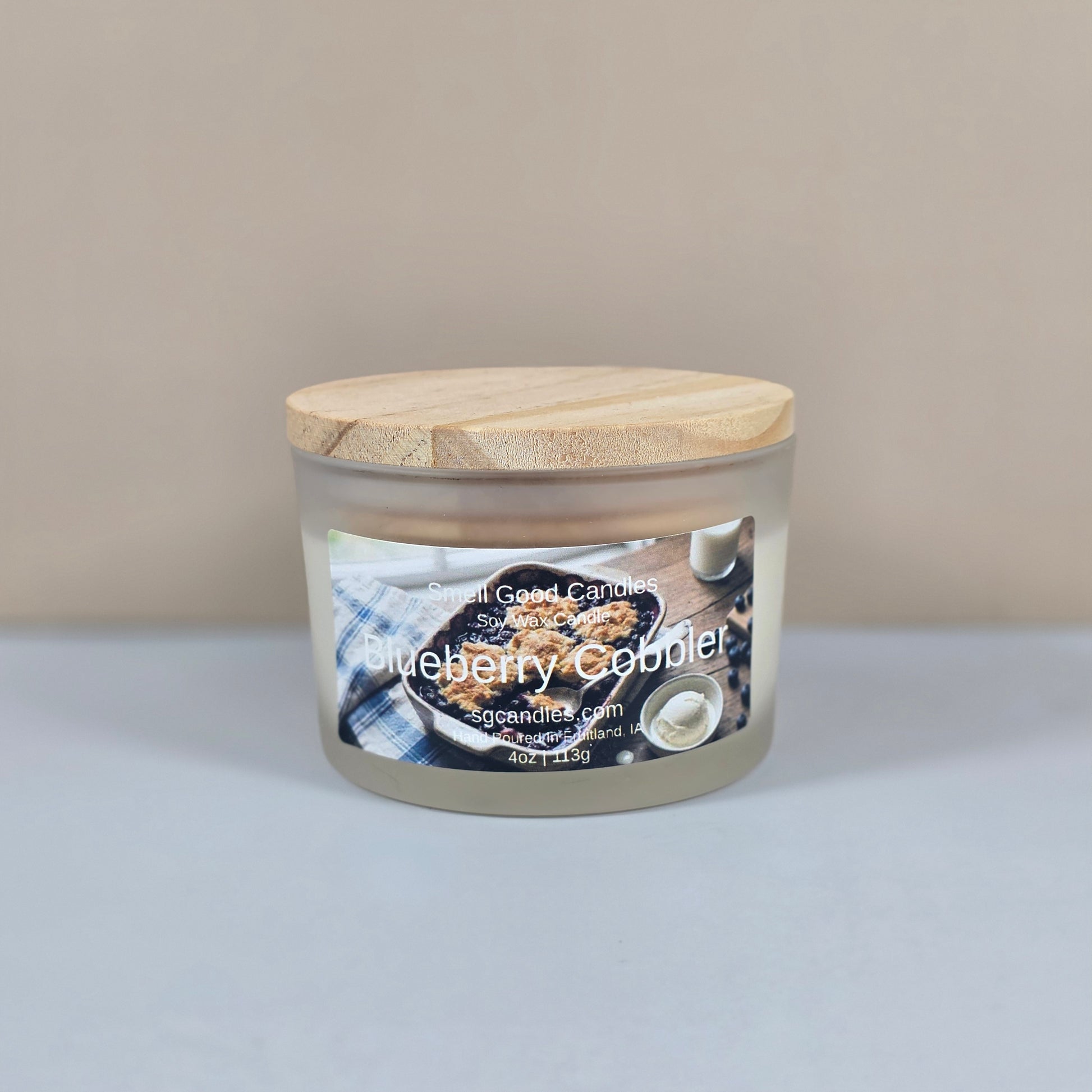 A 4oz clear glass jar candle with a black metal lid, featuring a "Blueberry Cobbler" label with a rustic illustration of a fresh blueberry dessert.