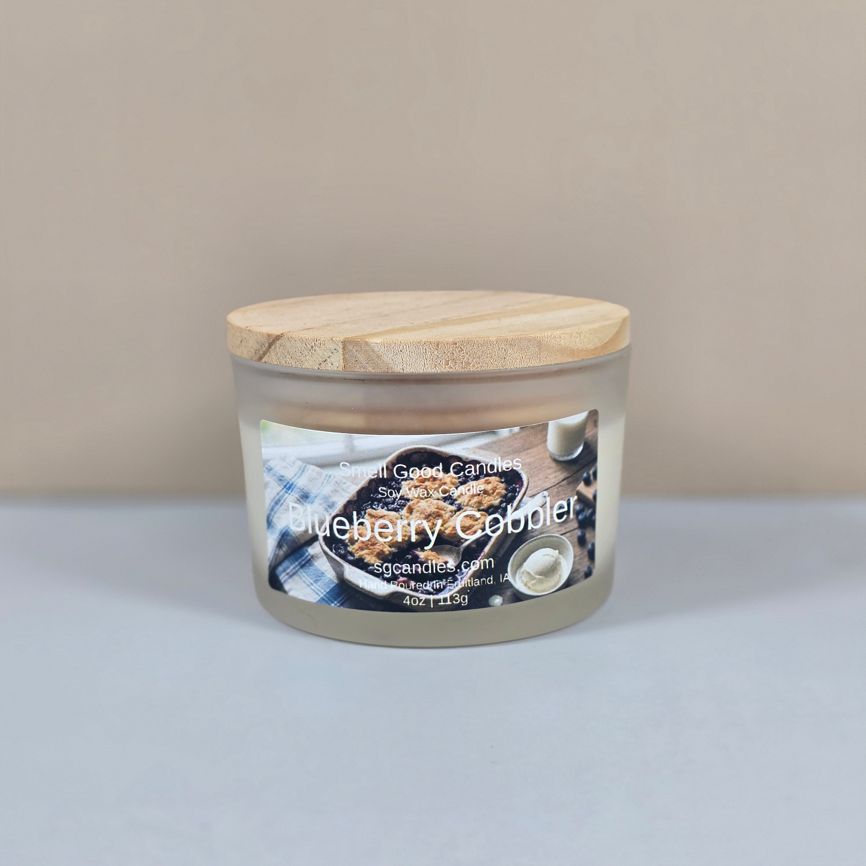 A 4oz clear glass jar candle with a black metal lid, featuring a "Blueberry Cobbler" label with a rustic illustration of a fresh blueberry dessert.