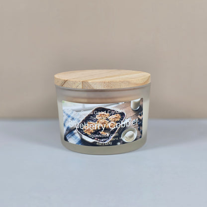 A 4oz clear glass jar candle with a black metal lid, featuring a "Blueberry Cobbler" label with a rustic illustration of a fresh blueberry dessert.