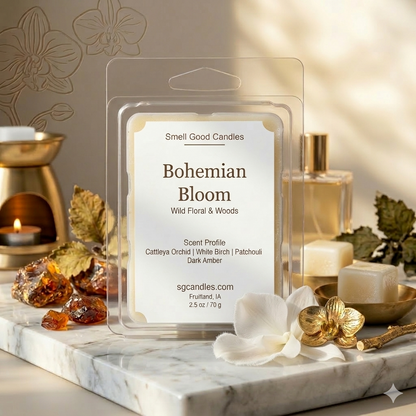 Smell Good Candles "Bohemian Bloom" Wild Floral & Woods Artisan Soy Wax Melt in a clear clamshell package on a luxury marble slab. Surrounding props include raw amber chunks, fresh Cattleya orchid petals, White Birch wood elements, and a stylized brass floral sculpture, lit by a warm, natural sunbeam.