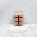 Pack of six tan-colored wax melts in clear packaging on a white marble surface with light gray background