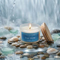 Lit "Cool Water" frosted glass jar candle from Small Good Candles in a serene aquatic flat lay setting with smooth river stones, sea glass, shells, and flowing water.