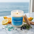 Luxury Cool Water soy wax candle in a white frosted glass jar with a natural wood lid and a realistic glowing flame, styled on a white marble surface with fragrance cues of fresh lemon, orange slices, melon, and aromatic herbs against a blurred coastal ocean background.