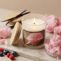 Handmade cotton candy soy wax candle in a white frosted glass jar with a natural wood lid, featuring a realistic glowing flame and surrounded by fragrance cues of pink spun sugar, fresh raspberries, blueberries, and vanilla beans on a white marble surface