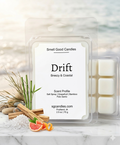 A luxury coastal-themed product photograph of "Drift" soy wax melts by Smell Good Candles. The minimalist white label features scent notes of salt spray, grapefruit, bamboo, and palo santo. The clear clamshell packaging is displayed on a white marble surface alongside fresh grapefruit slices, sea salt crystals, green bamboo stalks, and palo santo wood sticks. A soft-focus beach background with crashing waves and sunlight completes the breezy, high-end aesthetic.