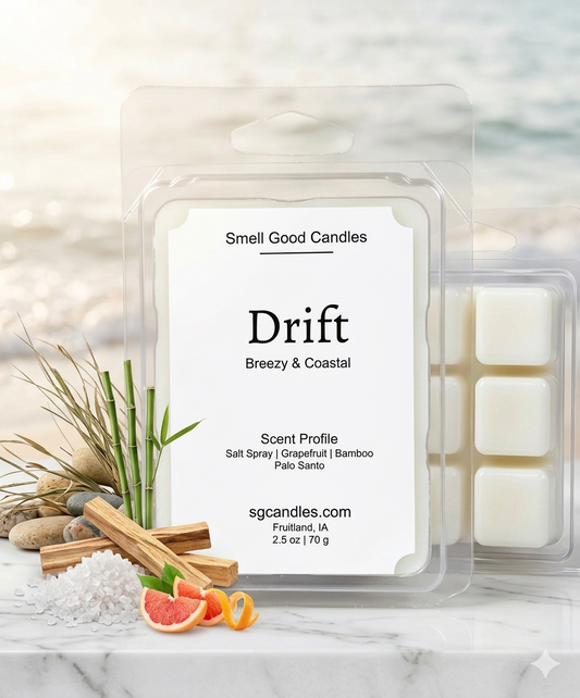 A luxury coastal-themed product photograph of "Drift" soy wax melts by Smell Good Candles. The minimalist white label features scent notes of salt spray, grapefruit, bamboo, and palo santo. The clear clamshell packaging is displayed on a white marble surface alongside fresh grapefruit slices, sea salt crystals, green bamboo stalks, and palo santo wood sticks. A soft-focus beach background with crashing waves and sunlight completes the breezy, high-end aesthetic.