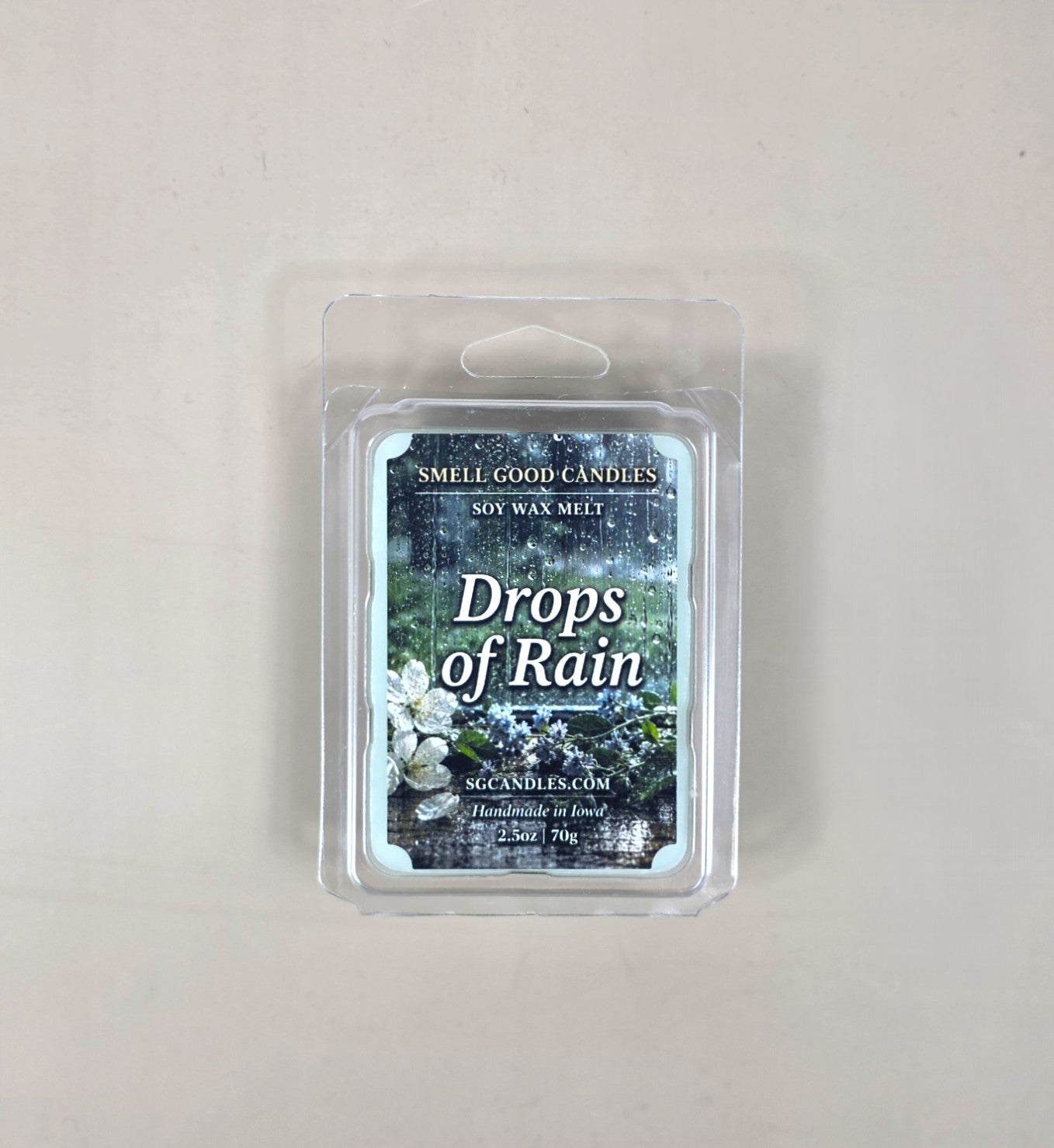 Top view of Drops of Rain fragrance soy wax melt in a 2.5oz clear clamshell package featuring a rainy window label design with fresh white and purple flowers.