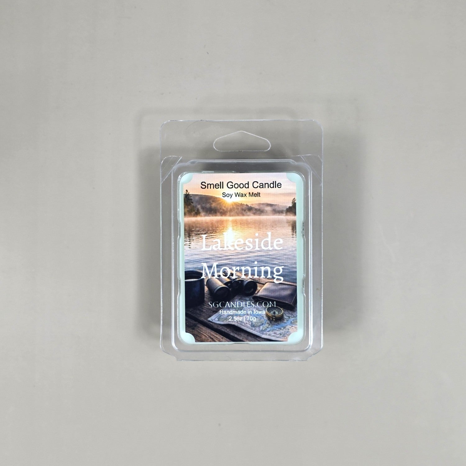 Labeled Lakeside Morning soy wax melt in a 2.5oz clamshell featuring a peaceful sunrise over a lake with binoculars and a map.