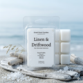A 2.5 oz clamshell of Smell Good Candles Linen & Driftwood wax melts resting on weathered coastal driftwood and fine sea sand, set against a calm ocean horizon on a crisp morning. The pure white wax cubes are visible through the clear plastic. The detailed white label features the product title, Sun-Drenched Cashmere description, and Scent Profile: Fresh Air, Cotton Blossom, Sea Salt, Driftwood. Sea salt crystals are scattered on the wood and sand.