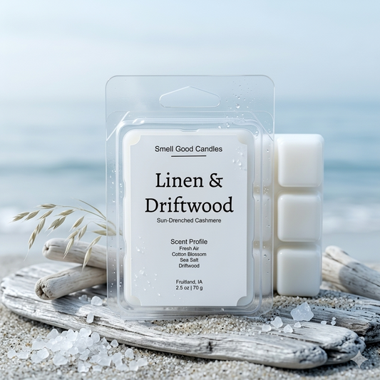 A 2.5 oz clamshell of Smell Good Candles Linen & Driftwood wax melts resting on weathered coastal driftwood and fine sea sand, set against a calm ocean horizon on a crisp morning. The pure white wax cubes are visible through the clear plastic. The detailed white label features the product title, Sun-Drenched Cashmere description, and Scent Profile: Fresh Air, Cotton Blossom, Sea Salt, Driftwood. Sea salt crystals are scattered on the wood and sand.