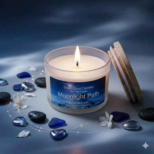 Lit "Moonlight Path" frosted glass candle by Smell Good Candles in a moody evening flat lay featuring white jasmine petals, polished black stones, and blue sea glass on a moonlit blue surface.