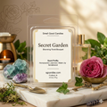 Artisan "Secret Garden" wax melt by Smell Good Candles in a clear 2.5 oz clamshell on a white marble slab. The luxury product photography includes a pink rose, green ivy, an antique brass key, a miniature brass trowel, and raw crystals under soft, dappled sunlight.