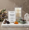 A professional product photograph of "Summit" soy wax melts by Smell Good Candles, featuring a minimalist label with scent notes of mountain air, black currant, cardamom, and amber. The clear clamshell packaging is displayed on a white marble slab alongside fresh black currants, cardamom pods, a piece of golden amber resin, and frosted pine branches. A clear quartz crystal and a bouquet of dried lavender and eucalyptus sit in the background against a moody, textured stone wall.