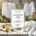 Smell Good Candles "Sun-Bleached Linen" artisan wax melt in a clear 2.5 oz clamshell, featured on a weathered white wooden surface. The scene includes pure white wax cubes, fresh lemon zest in a small glass bowl, and white birch logs to represent the scent profile of Lemon Zest, Water Lily, White Birch, and Soft Musk. In the blurred background, clean white linens hang from a natural jute clothesline under bright, direct morning sunlight.
